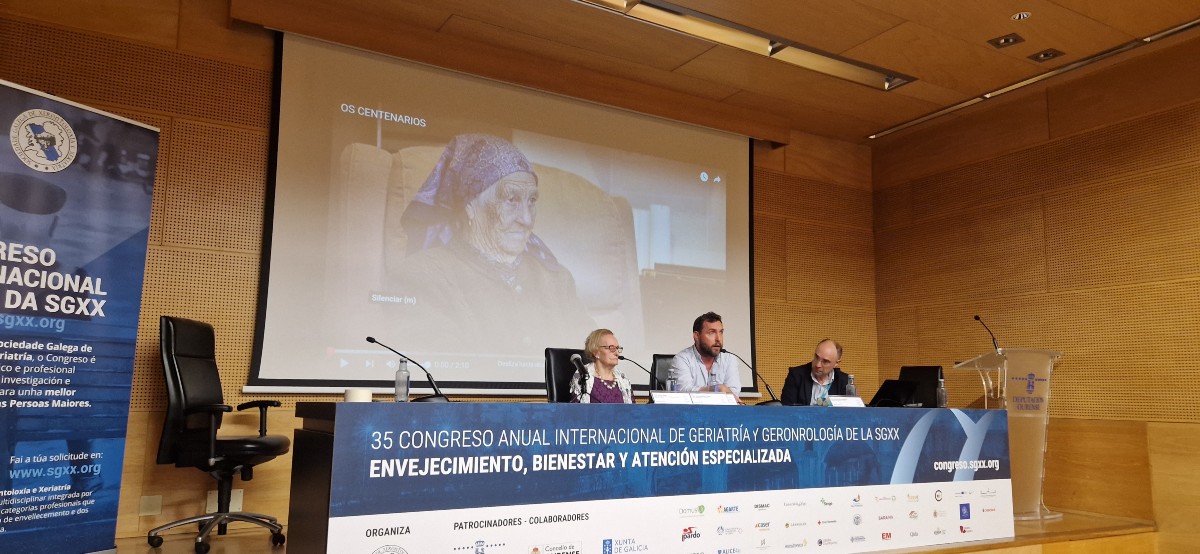 Rubén Riós presenta el documental “Centenar10s” en el 35º Congreso de la SGXX, con una imagen proyectada de una mujer centenaria y el mensaje “Os centenarios”.