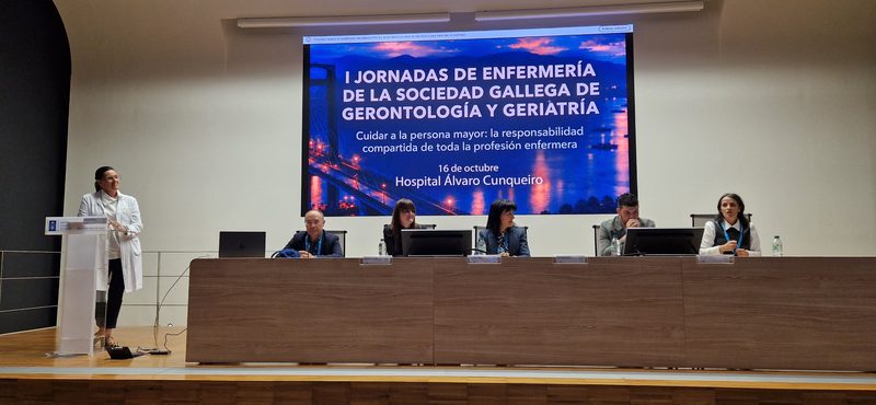 Mesa de inauguración de la jornada en el escenario del Hospital Álvaro Cunqueiro con el cartel de las I Jornadas de Enfermería de la Sociedad Gallega de Gerontología y Geriatría; una ponente interviene desde el atril