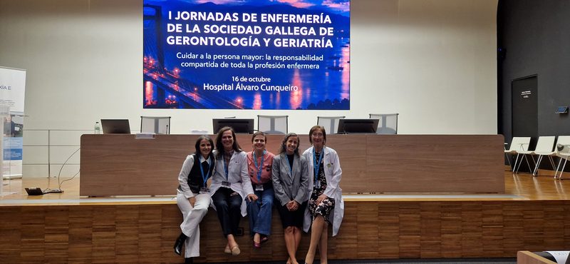 Cinco profesionales posan juntas sobre el escenario del salón de actos, con el cartel de las I Jornadas de Enfermería de la Sociedad Gallega de Gerontología y Geriatría en la pantalla